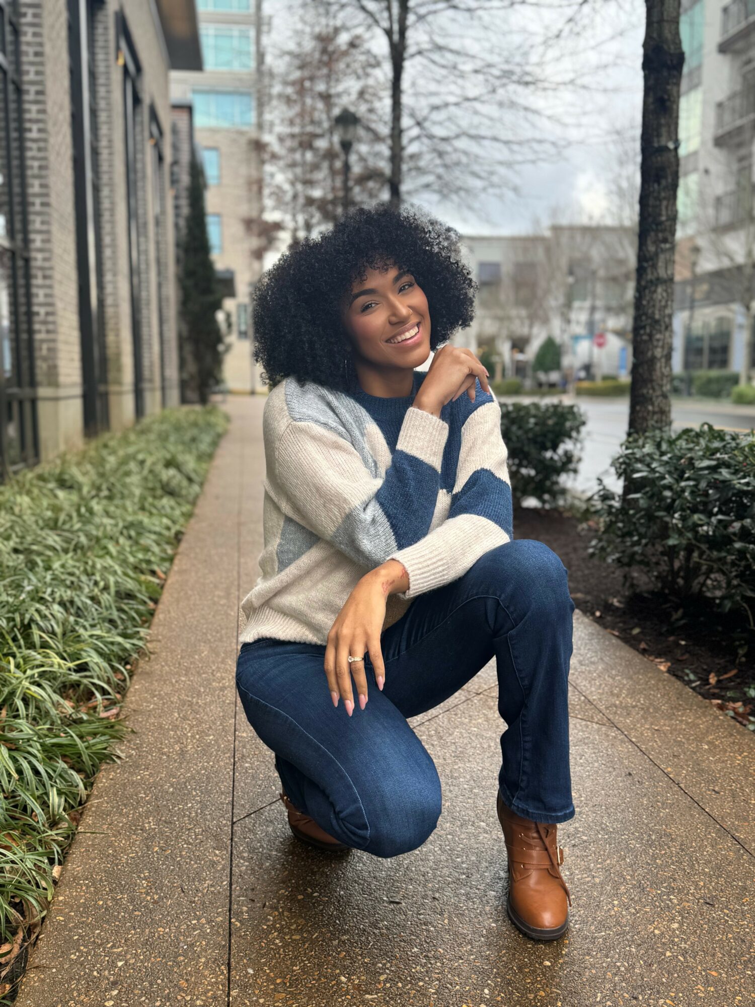 Young woman with curly hair crouching on a sidewalk in an urban setting, smiling, wearing a striped sweater, jeans, and brown boots.