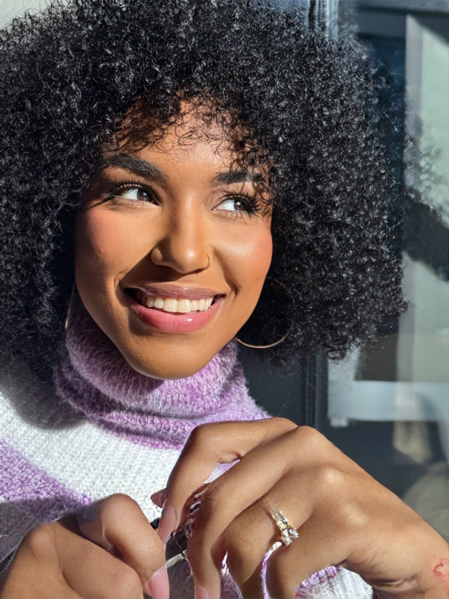Smiling woman with curly hair wearing a purple scarf, sitting near a window with sunlight, holding her hands near her face.