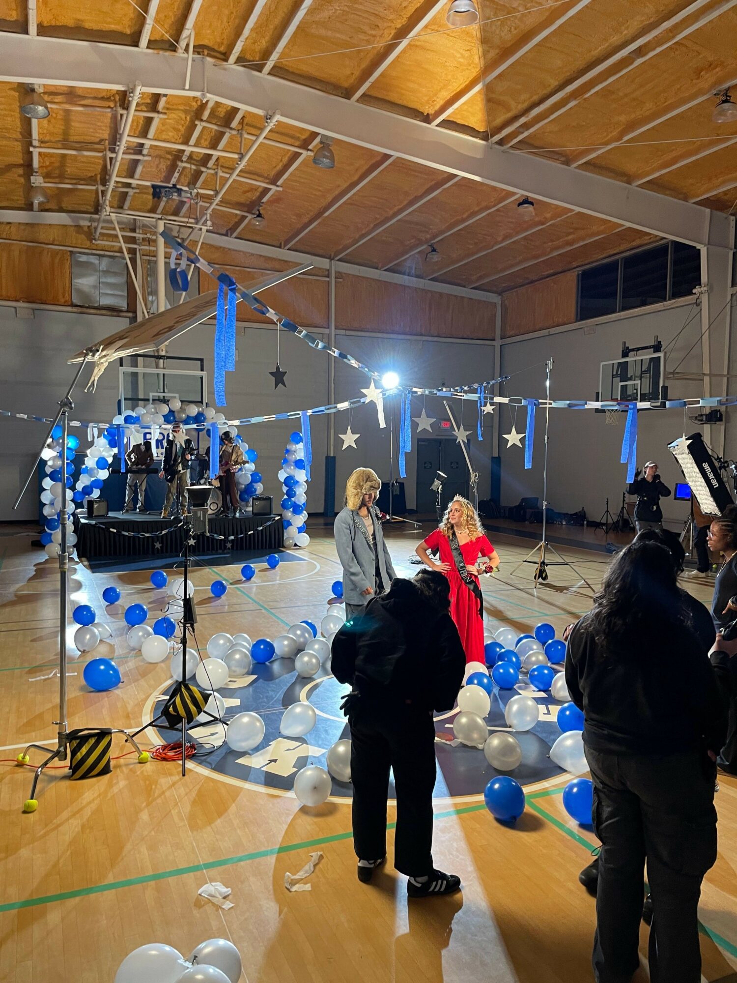 People in a gymnasium decorated with blue and white balloons and streamers, with a table and a stage in the background.