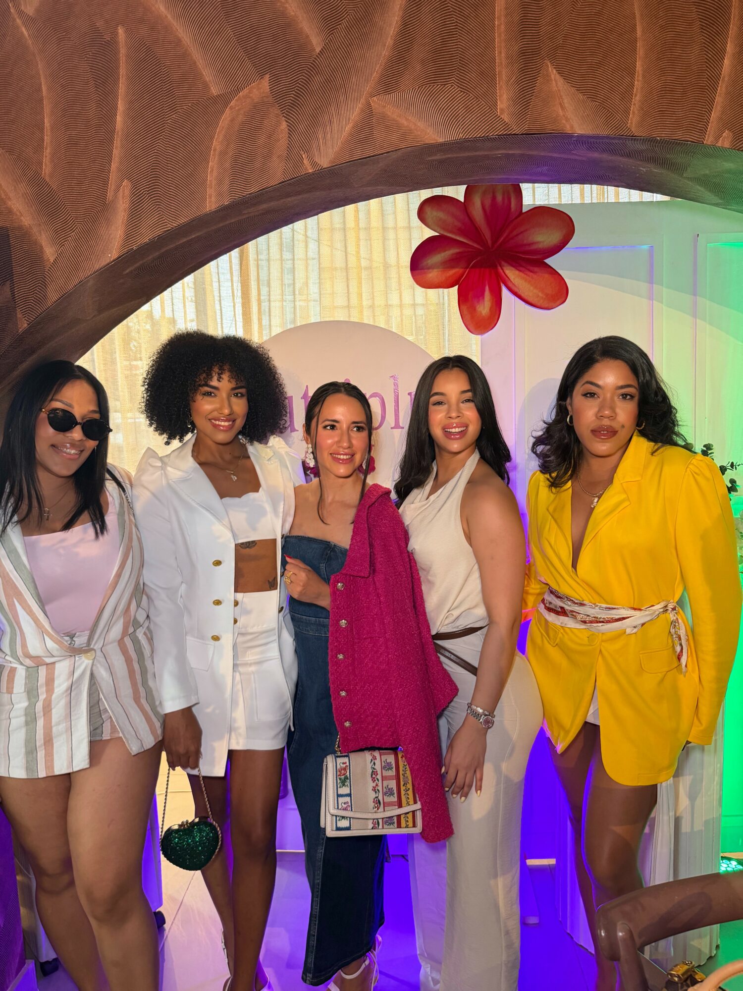 Five women standing together in colorful outfits, smiling, with a rainbow background and a large red flower decoration.