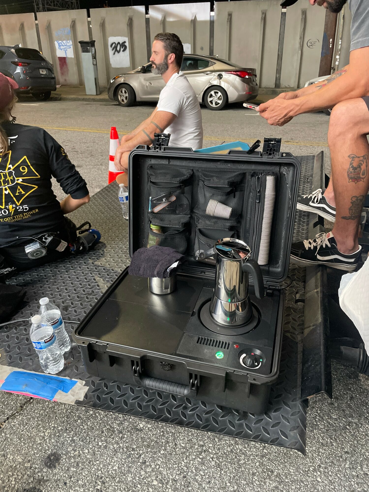 Portable device with a kettle, placed on a black case, with people sitting and standing nearby in an outdoor parking area.