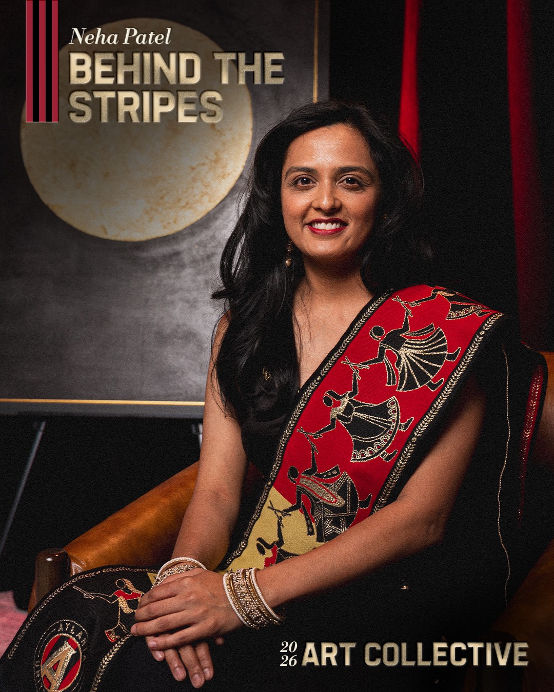 Woman in black dress with red and gold patterned shawl, sitting on a chair, smiling, in front of a poster for 'Behind the Stripes' exhibition.