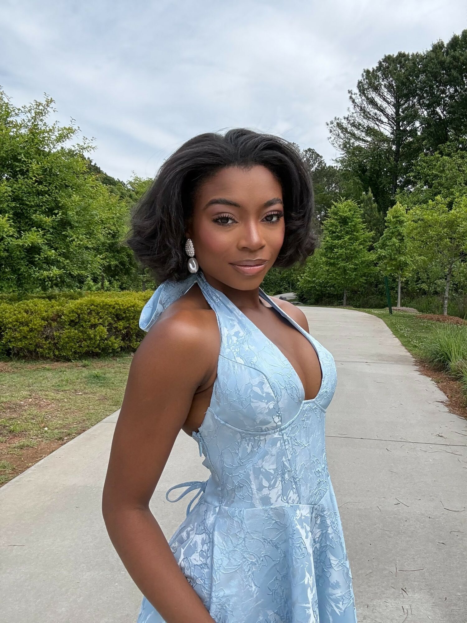 Young woman with shoulder-length black hair in a light blue dress outdoors, trees and a pathway in the background.