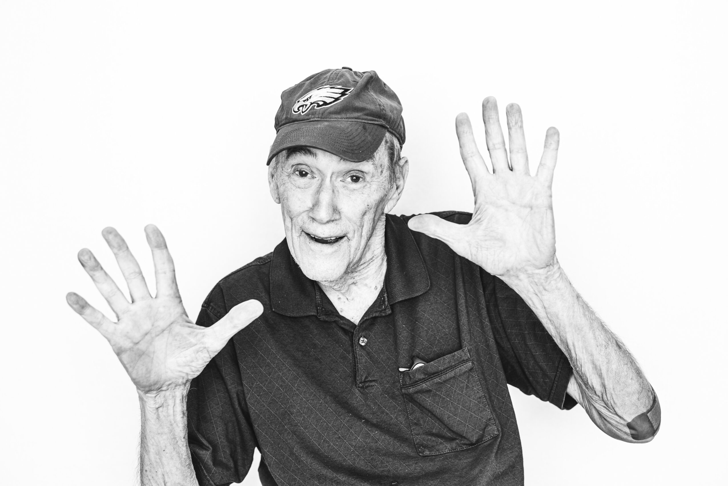 Older person with short hair wearing a cap and dark shirt, holding hands up with palms facing forward, smiling.