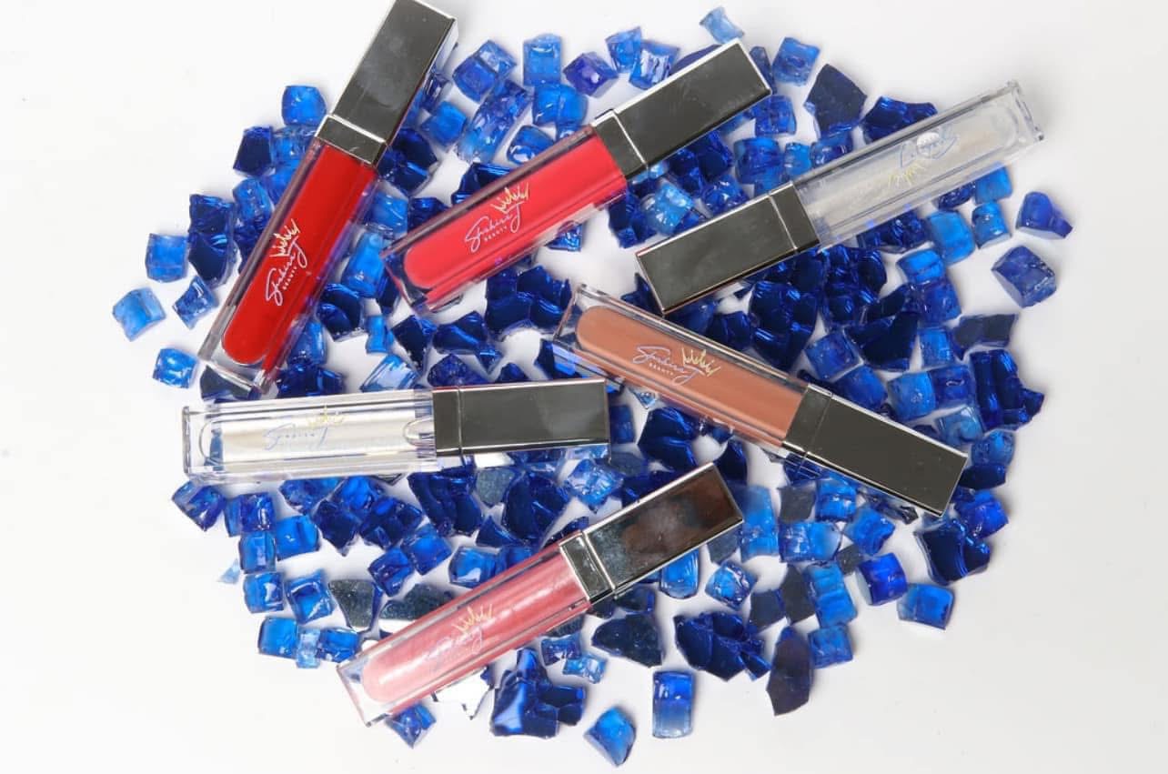 Lip gloss tubes placed on blue mosaic tiles, with some tubes overlapping, in various shades of red, pink, and nude.