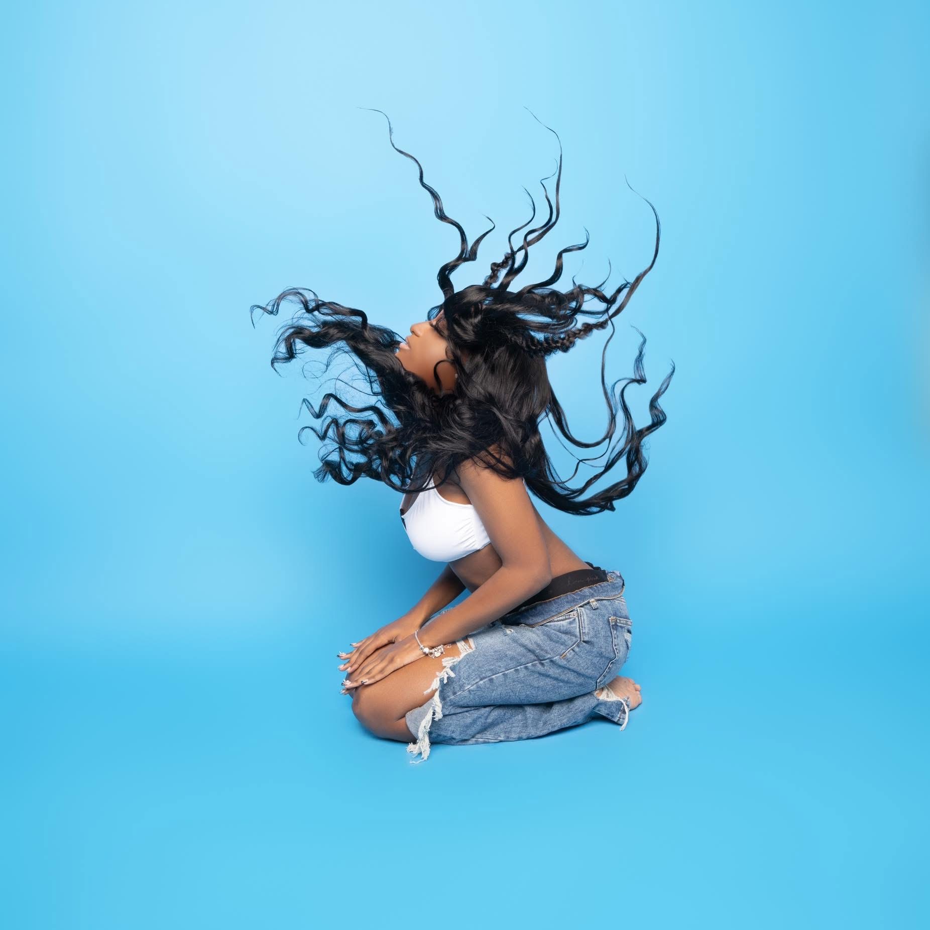 Person with long hair in motion, kneeling against a blue background, hair flying around, wearing a white top and denim shorts.