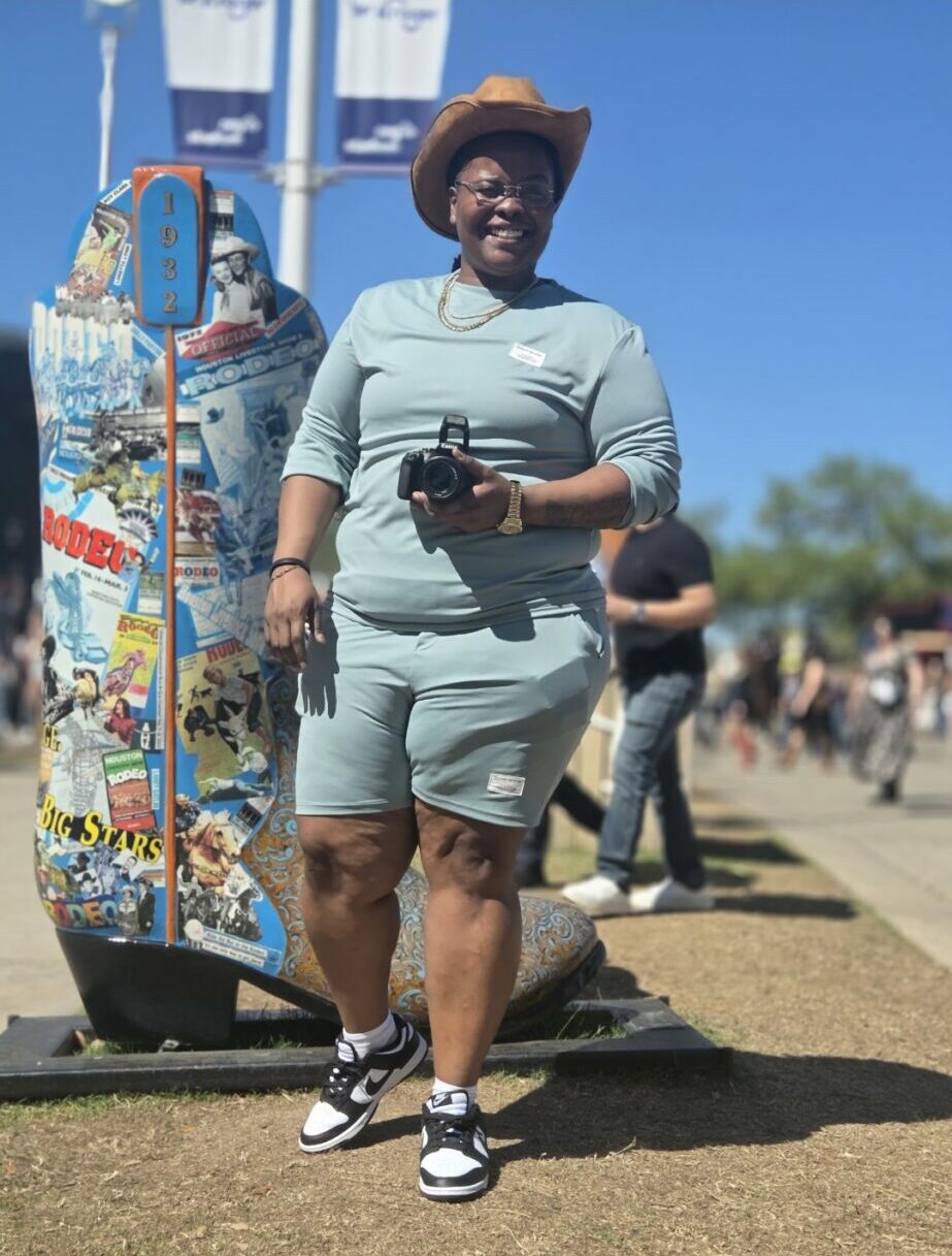 Person walking outdoors holding a skateboard, wearing a hat, sunglasses, and casual clothing, with a clear blue sky in background.