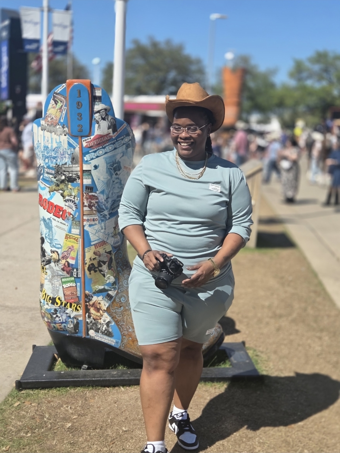 Smiling woman in light blue outfit and cowboy hat standing outdoors near a skateboard display, holding gloves, with people in background.