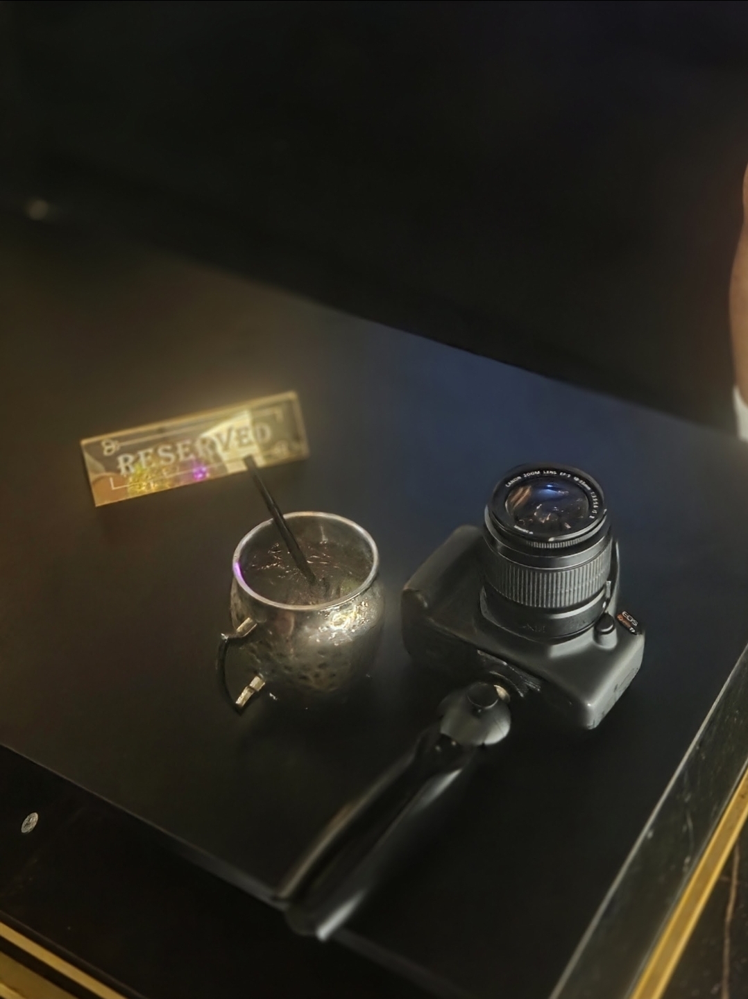 Camera, glass with straw, and a sign on a dark surface, viewed from above.