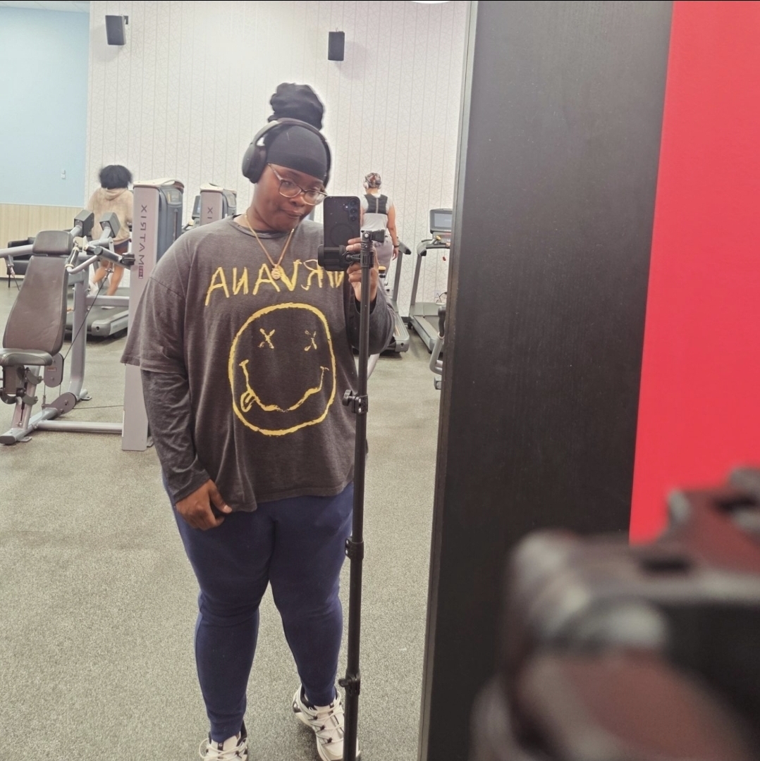 Person taking a mirror selfie in a gym, wearing headphones, a smiley face shirt, and dark pants.