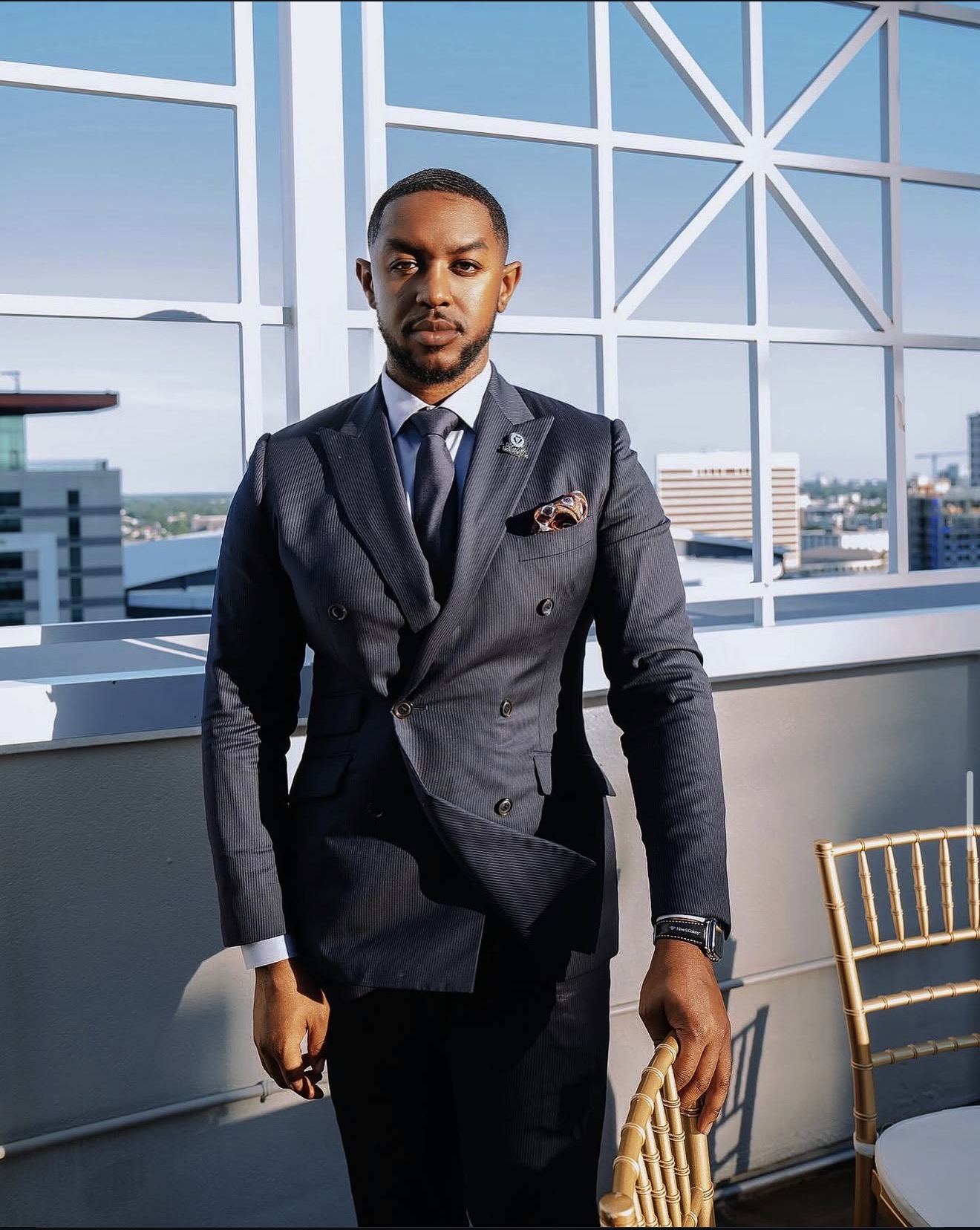 Man in a dark suit standing indoors with large windows and cityscape view behind him.