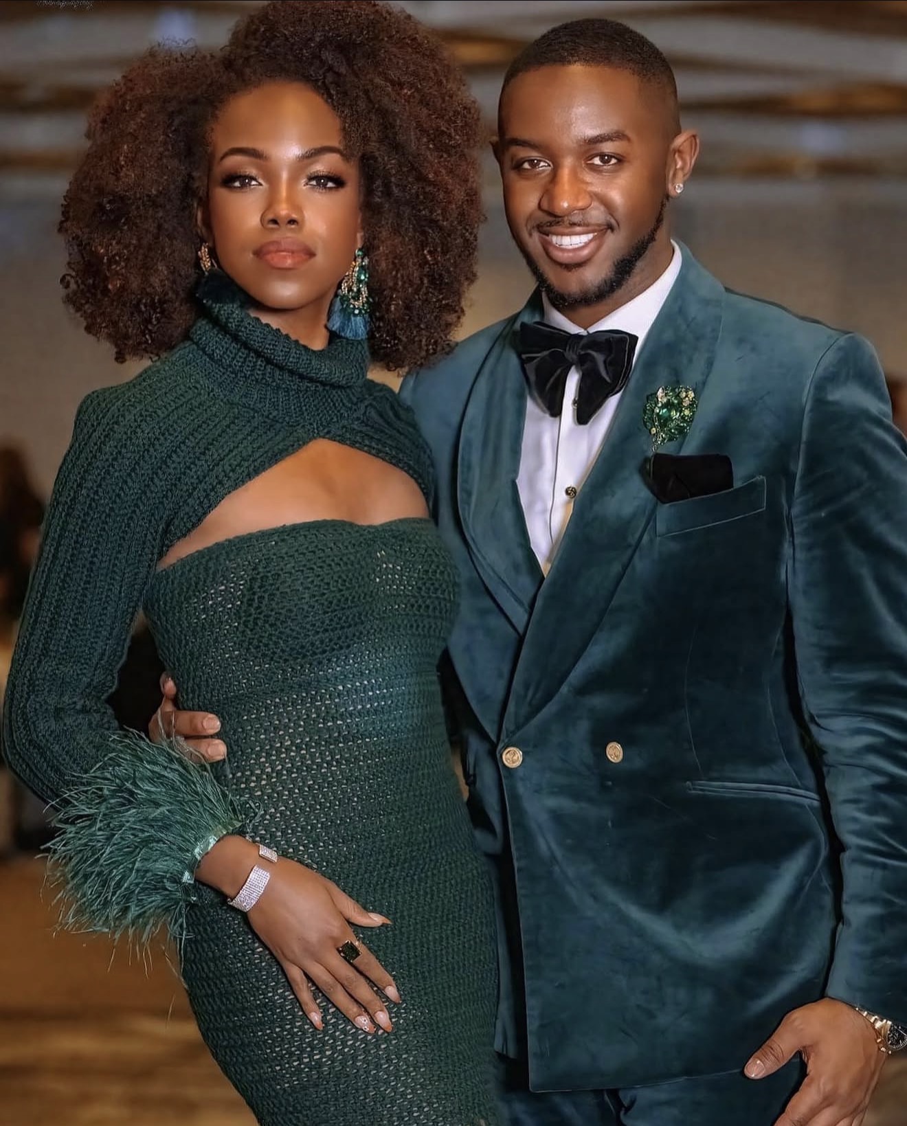 A woman and man dressed formally, standing close together, smiling, with a blurred background.