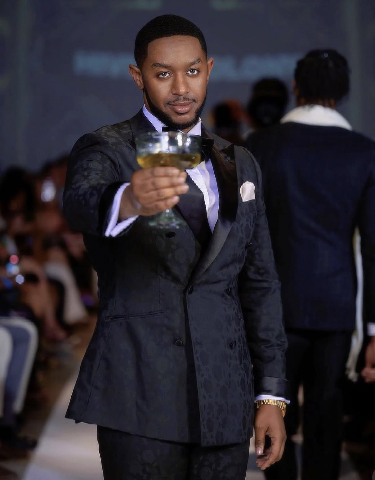 Man in tuxedo holding a glass, at a formal event, with blurred background of people and dark setting.