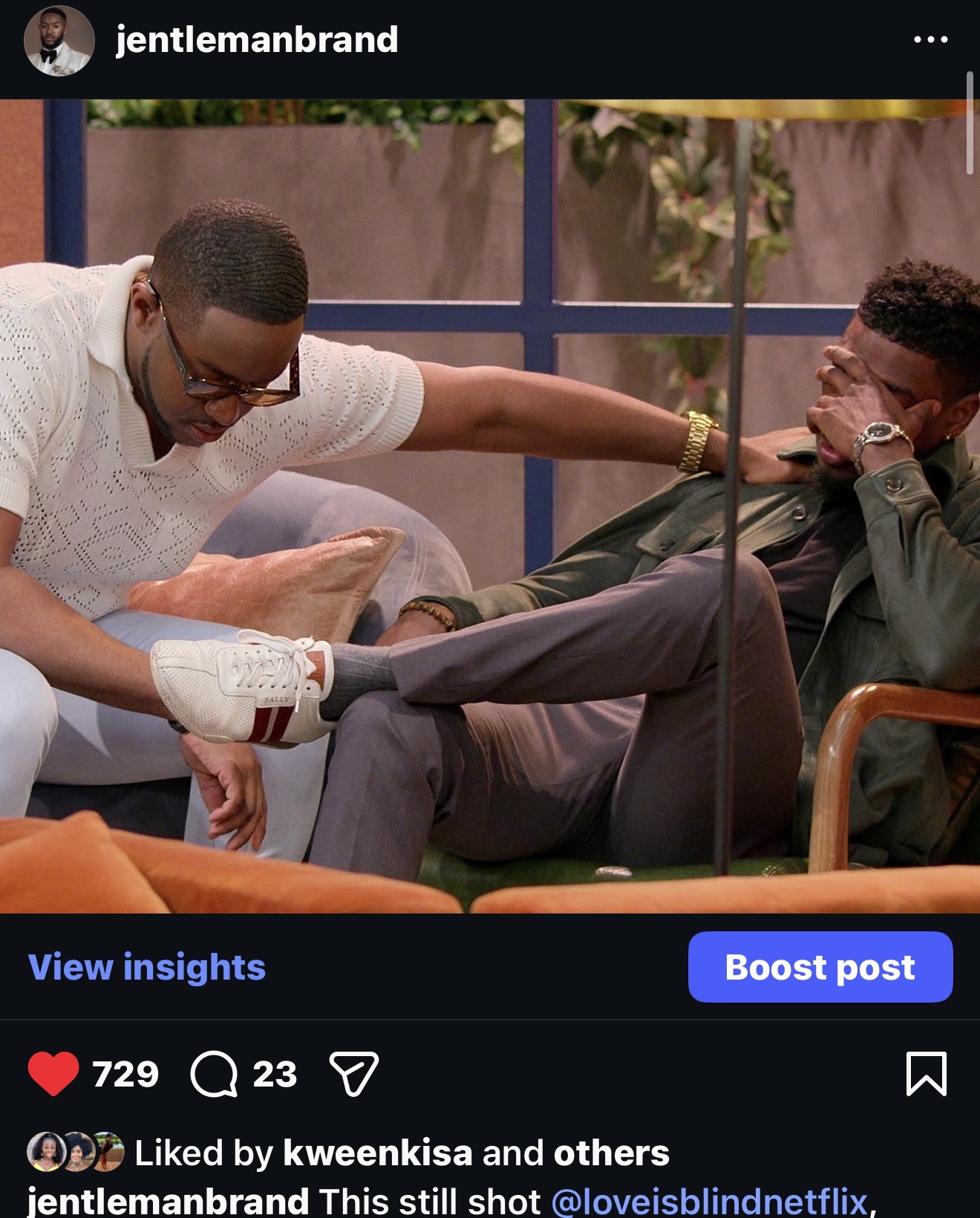 Two men are sitting on a couch, one is reaching out and touching the other's leg, both are smiling and laughing.