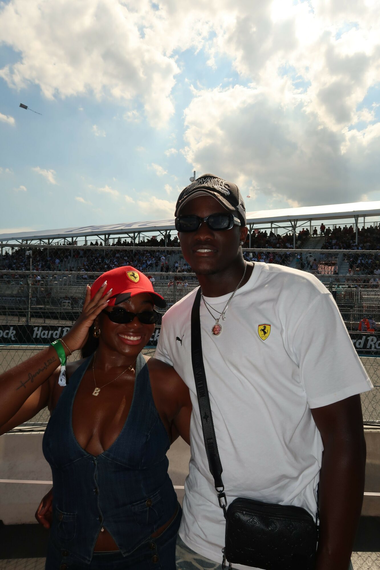 Two people outdoors, one wearing sunglasses and a white shirt, the other in a red cap, with a cloudy sky and water in background.