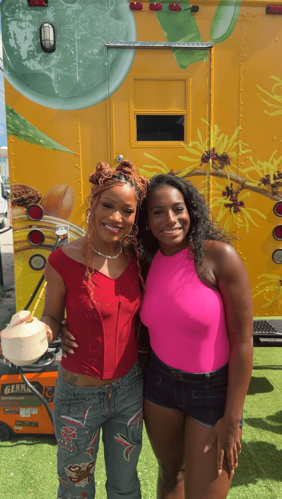 Two women smiling, one in a red top and jeans, the other in a pink top and black shorts, standing outdoors.
