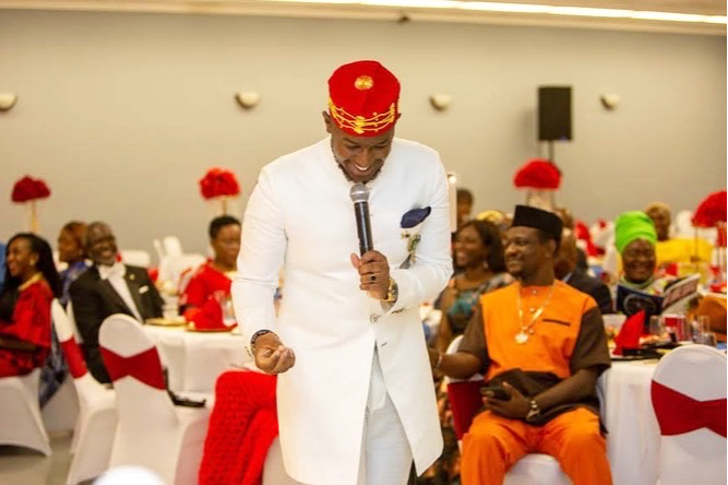 Man in white traditional attire and red hat speaking into a microphone at a celebration with seated guests and red decorations