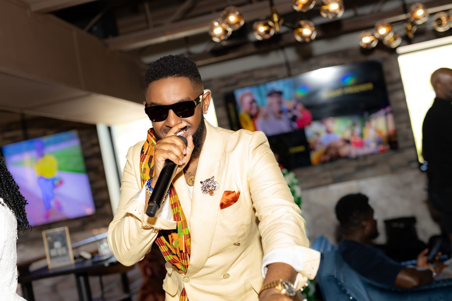 Man wearing sunglasses and a cream-colored suit singing into a microphone in a lively indoor setting.