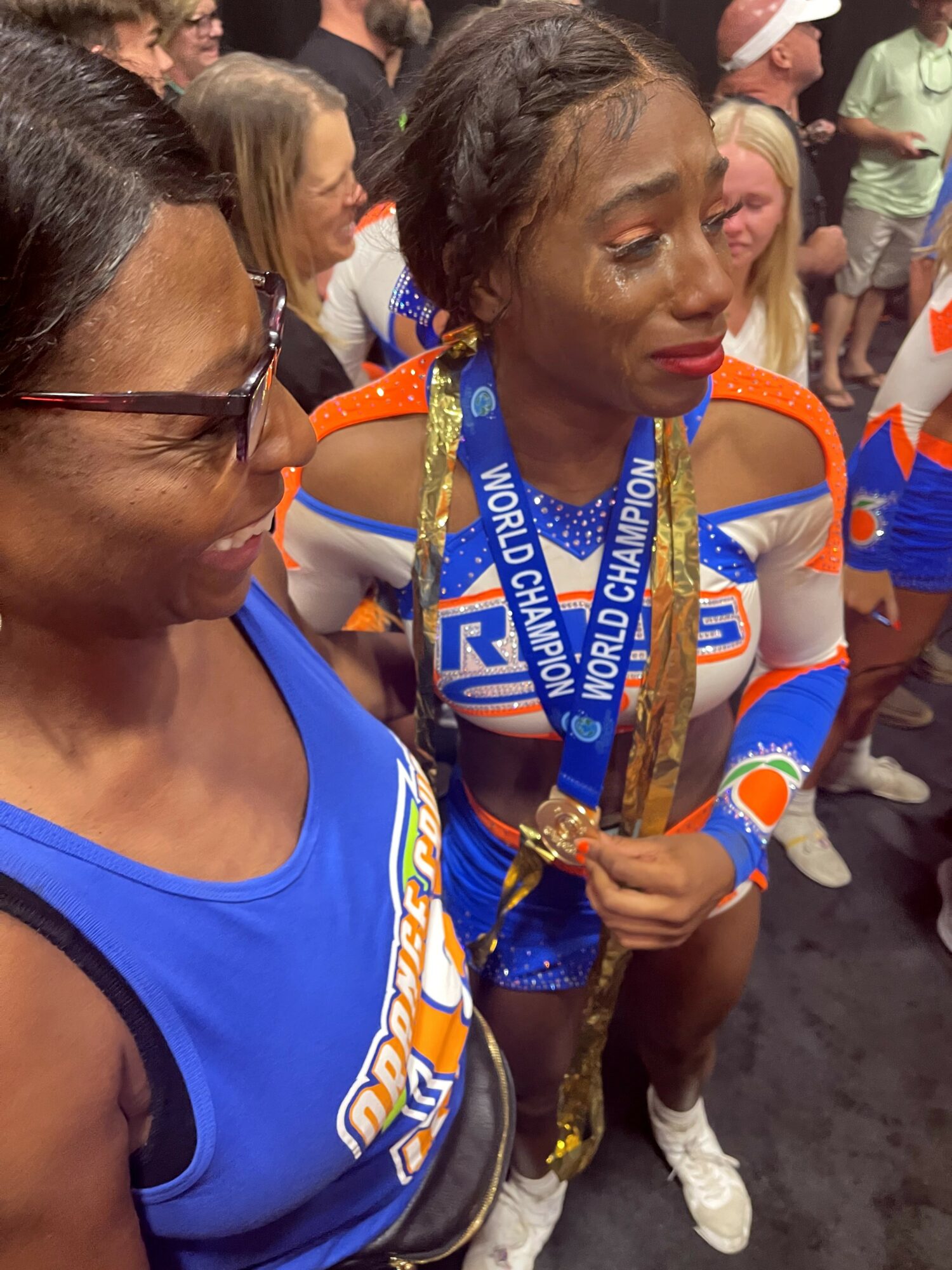 Two women in blue sports uniforms with medals, surrounded by people, celebrating a victory, one woman holding a gold trophy.