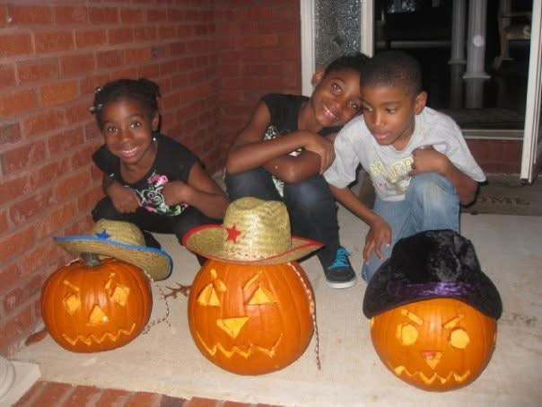 Three children sit on the ground with three decorated pumpkins, two wearing hats, in front of a brick wall and door.