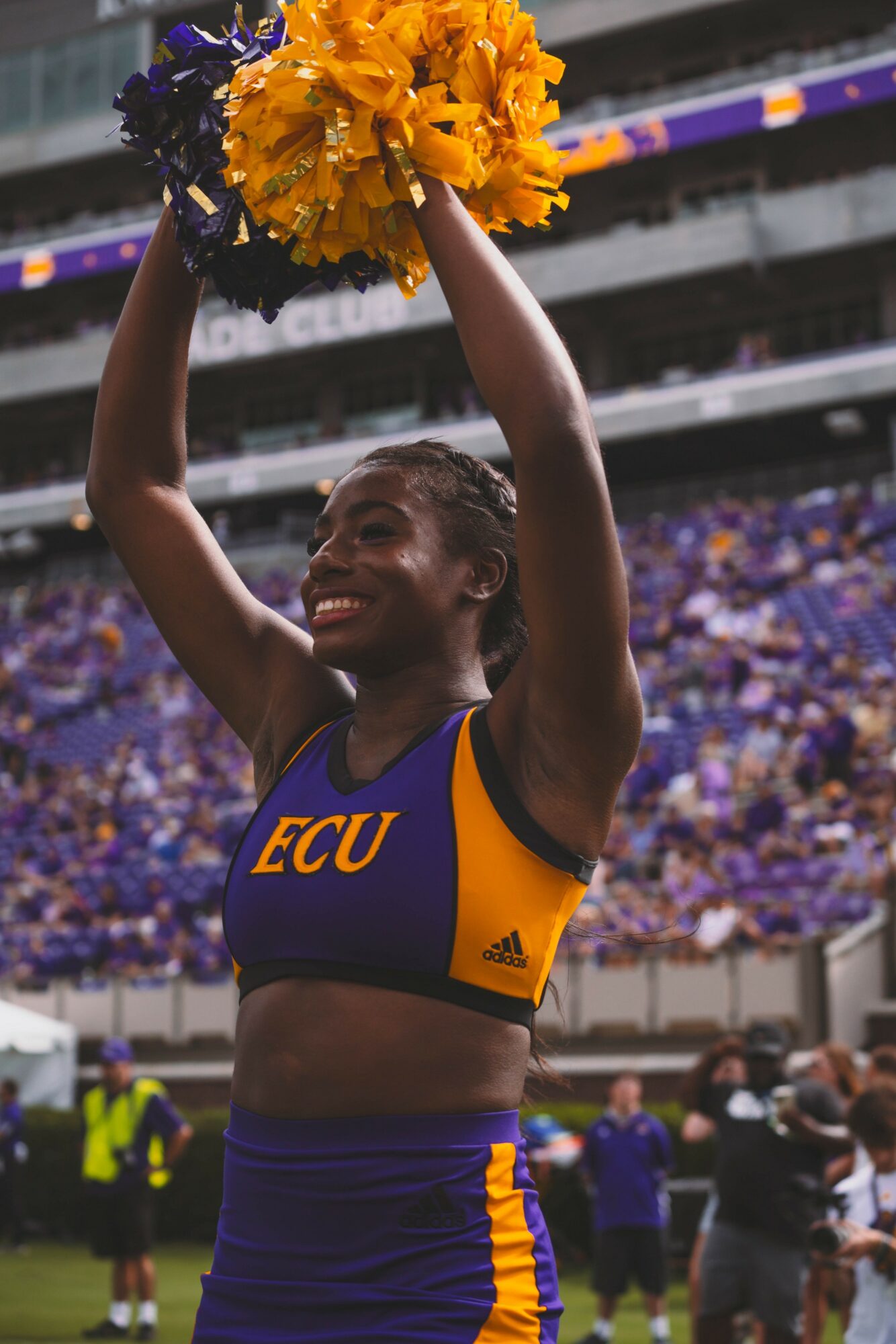 Cheerleader holding yellow pom-poms above head, smiling, in a stadium with purple seats and crowd, wearing ECU uniform.