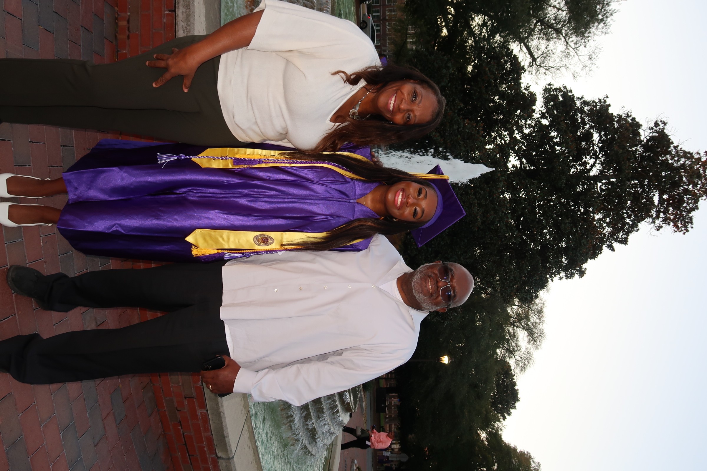Three people standing outdoors, one in a graduation gown and cap, two others in casual and formal attire, trees in background.
