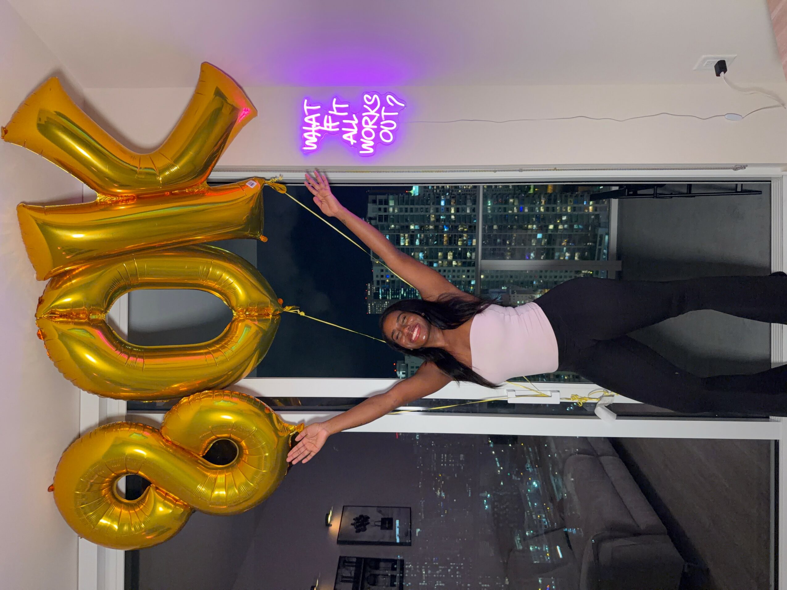Person holding large gold balloons shaped as the number 18, standing in front of a window with city view at night.