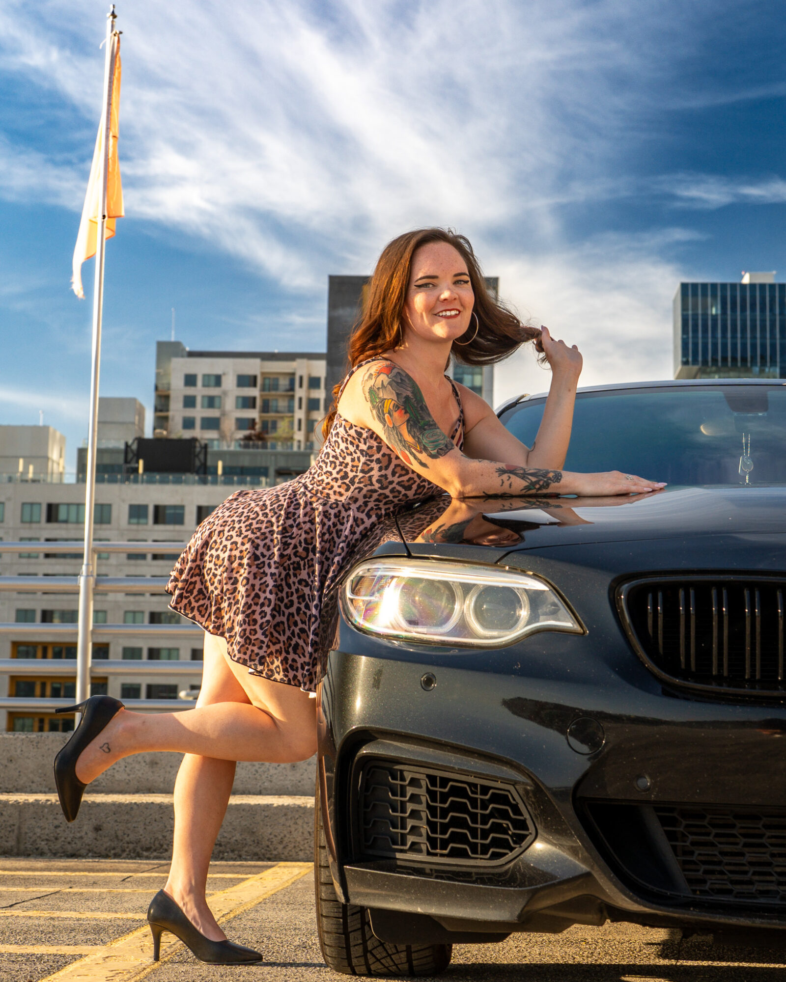 Smiling woman with tattoos leaning on a black car in an urban setting with buildings and a flagpole, sunny sky.