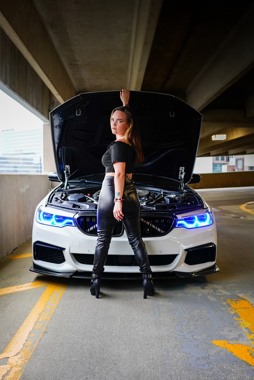 Woman standing in front of a car with open hood, under a parking garage ceiling, with city buildings in background.