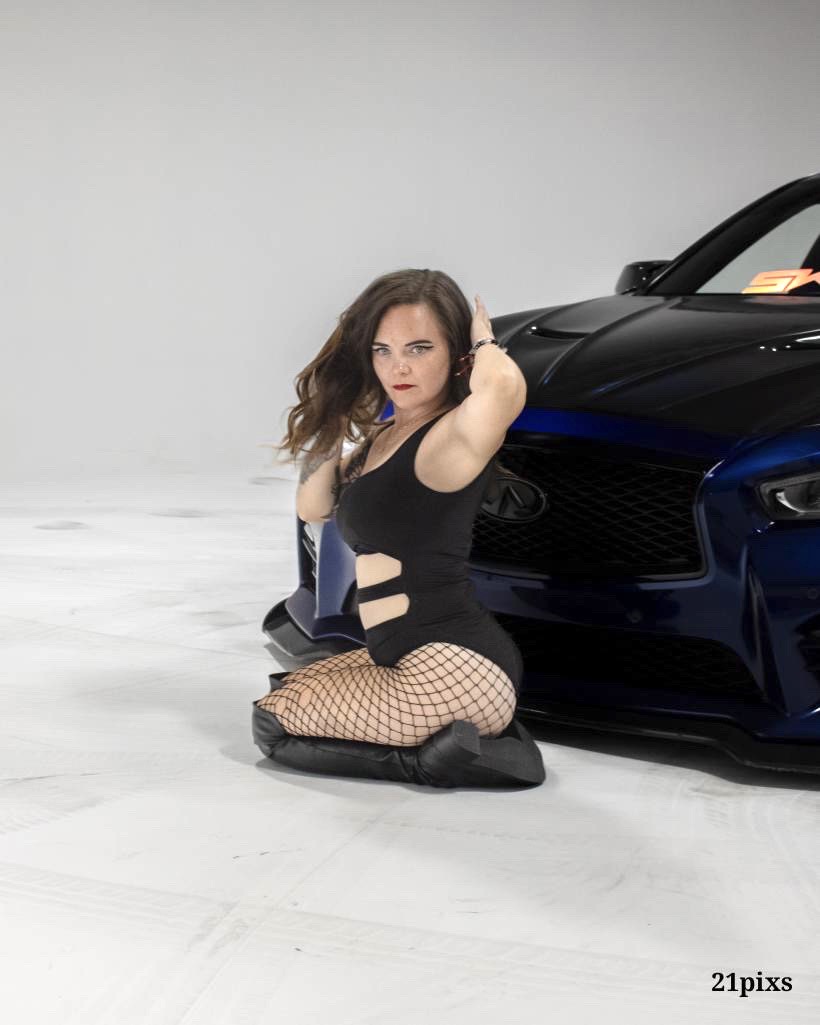 Woman kneeling next to a black sports car, wearing a black dress with cutouts and fishnet stockings.