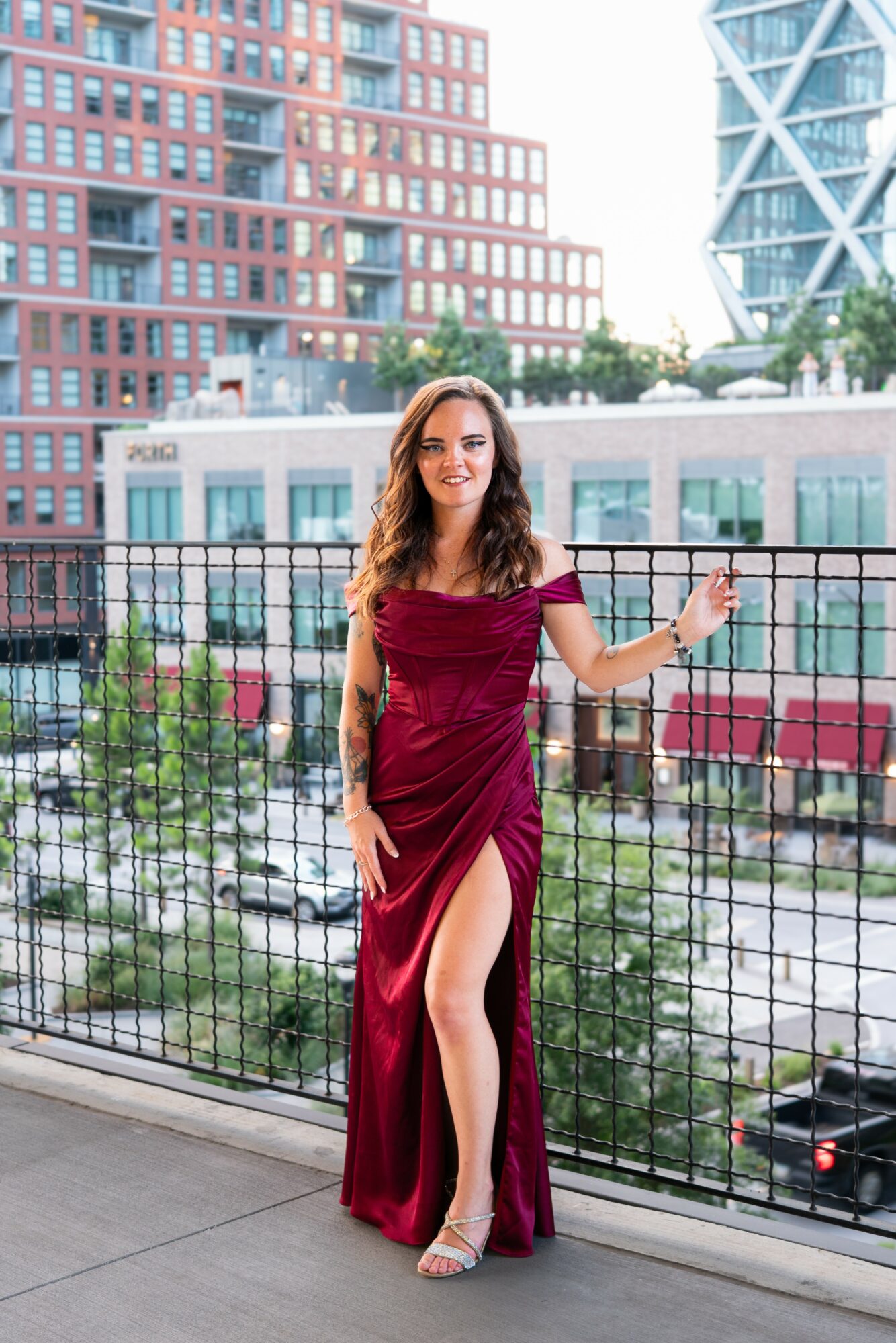 Woman in a burgundy dress with a high slit standing on a balcony with city buildings in background.