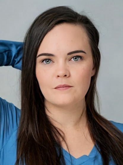 Portrait of a woman with long dark hair, blue eyes, and a neutral expression, wearing a blue top, against a light background.
