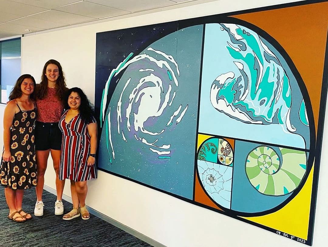 Three women stand next to a large circular artwork depicting swirling ocean waves and smaller circular designs, on a white wall.