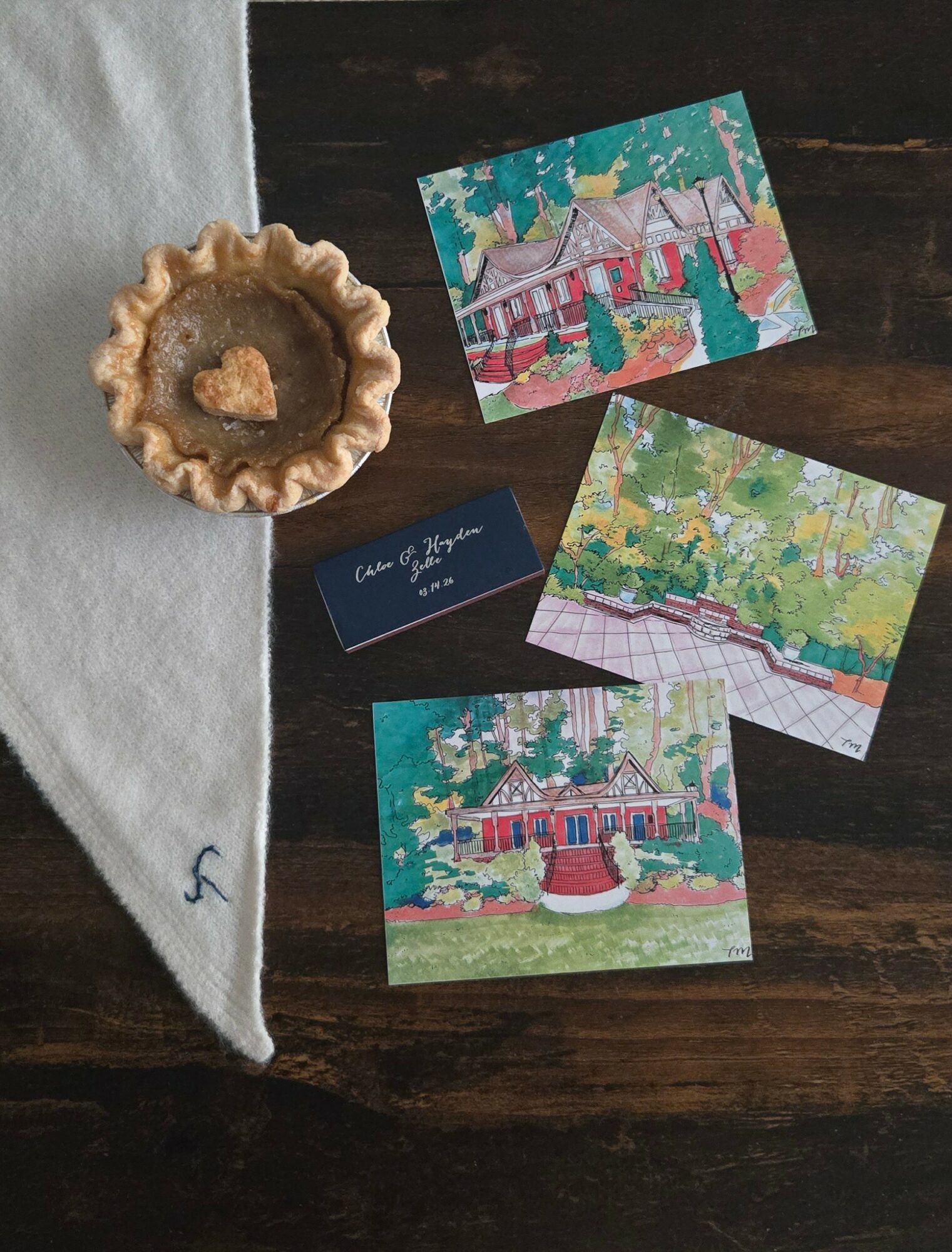 Three colorful illustrated postcards, a small tart with a heart-shaped filling, and a dark blue card on a wooden surface.