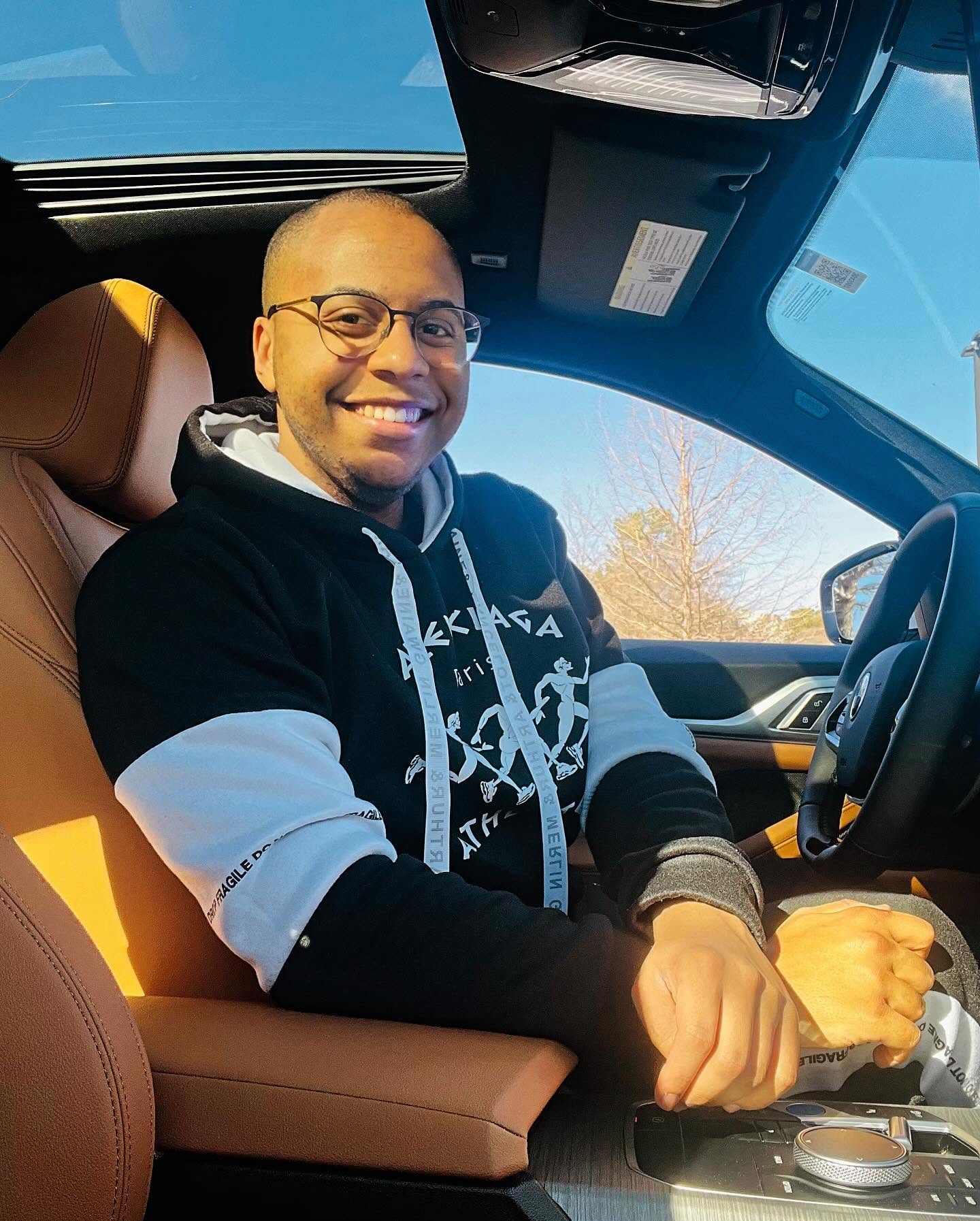 Smiling person with glasses sitting in a car, wearing a black and white hoodie, with a view of the sky outside.