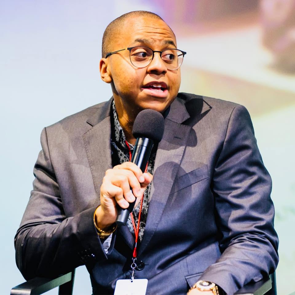 Person speaking into a microphone, wearing glasses and a suit, with a blurred background.