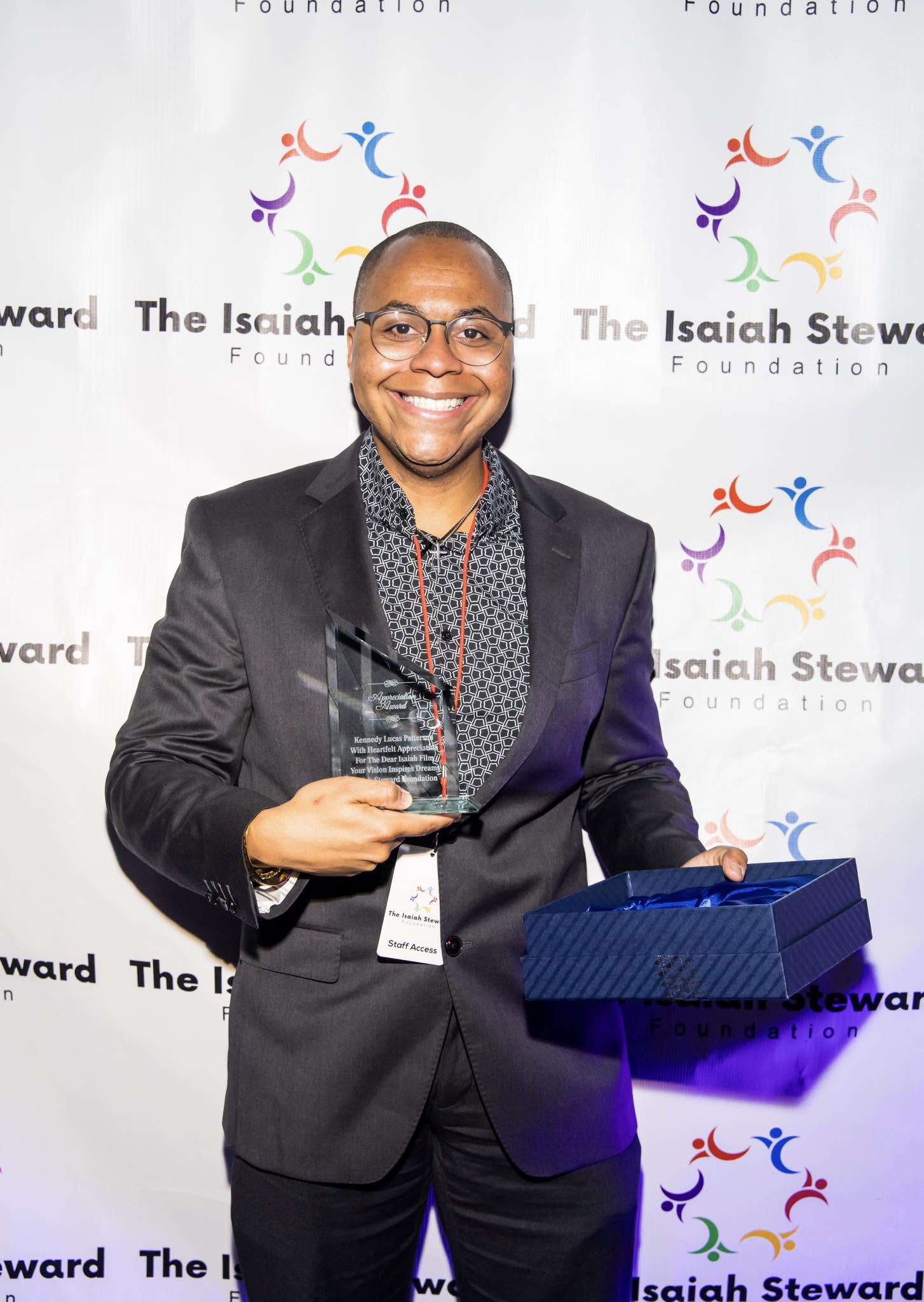 Person smiling, holding an award and a blue box, standing in front of a backdrop with colorful logo and text.