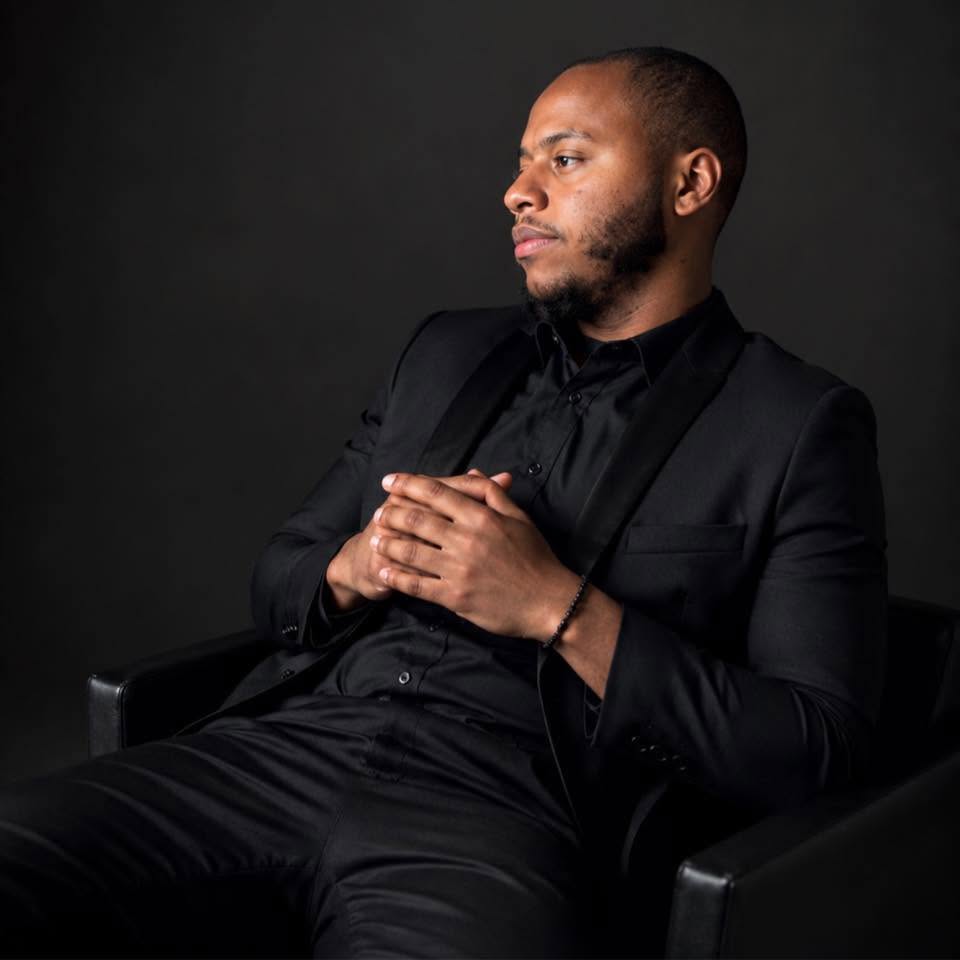 Man in black suit sitting with hands clasped, looking to the side against dark background.
