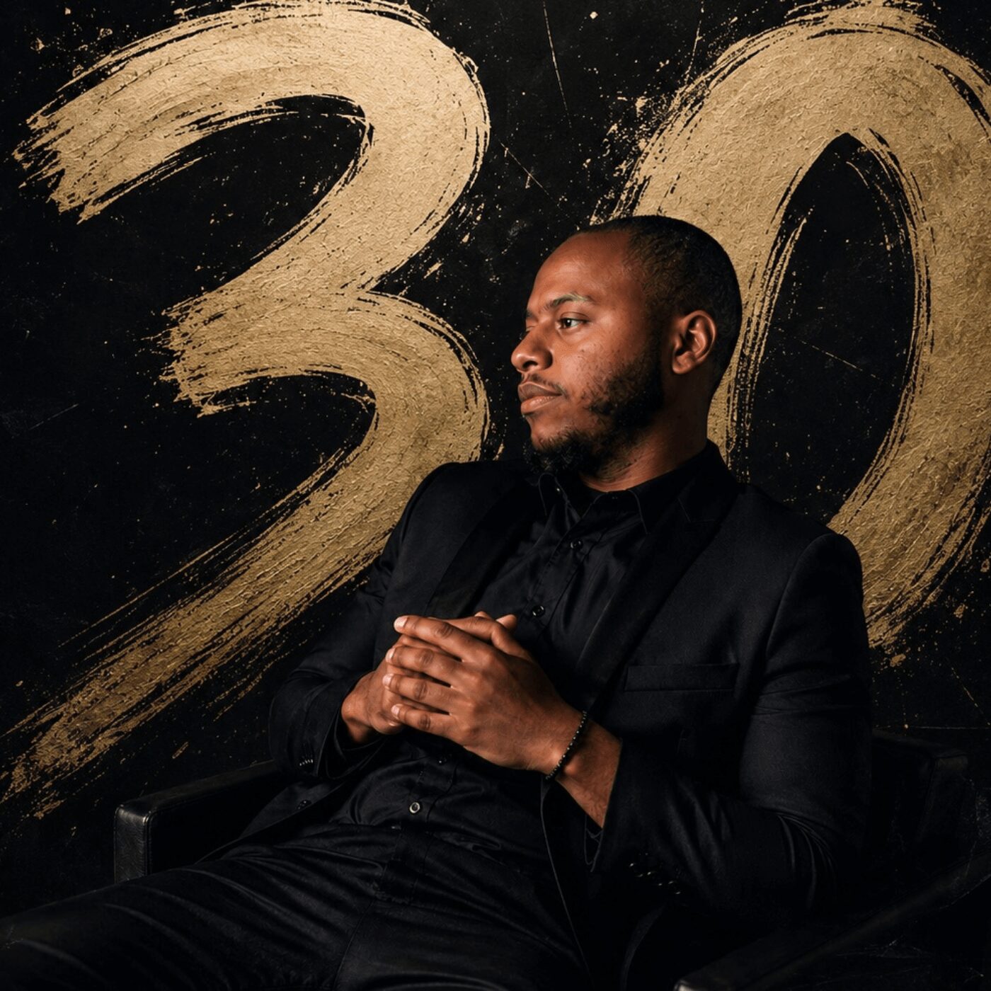 Man in black suit sitting with hands clasped, background with large gold number 30, looking to the side.