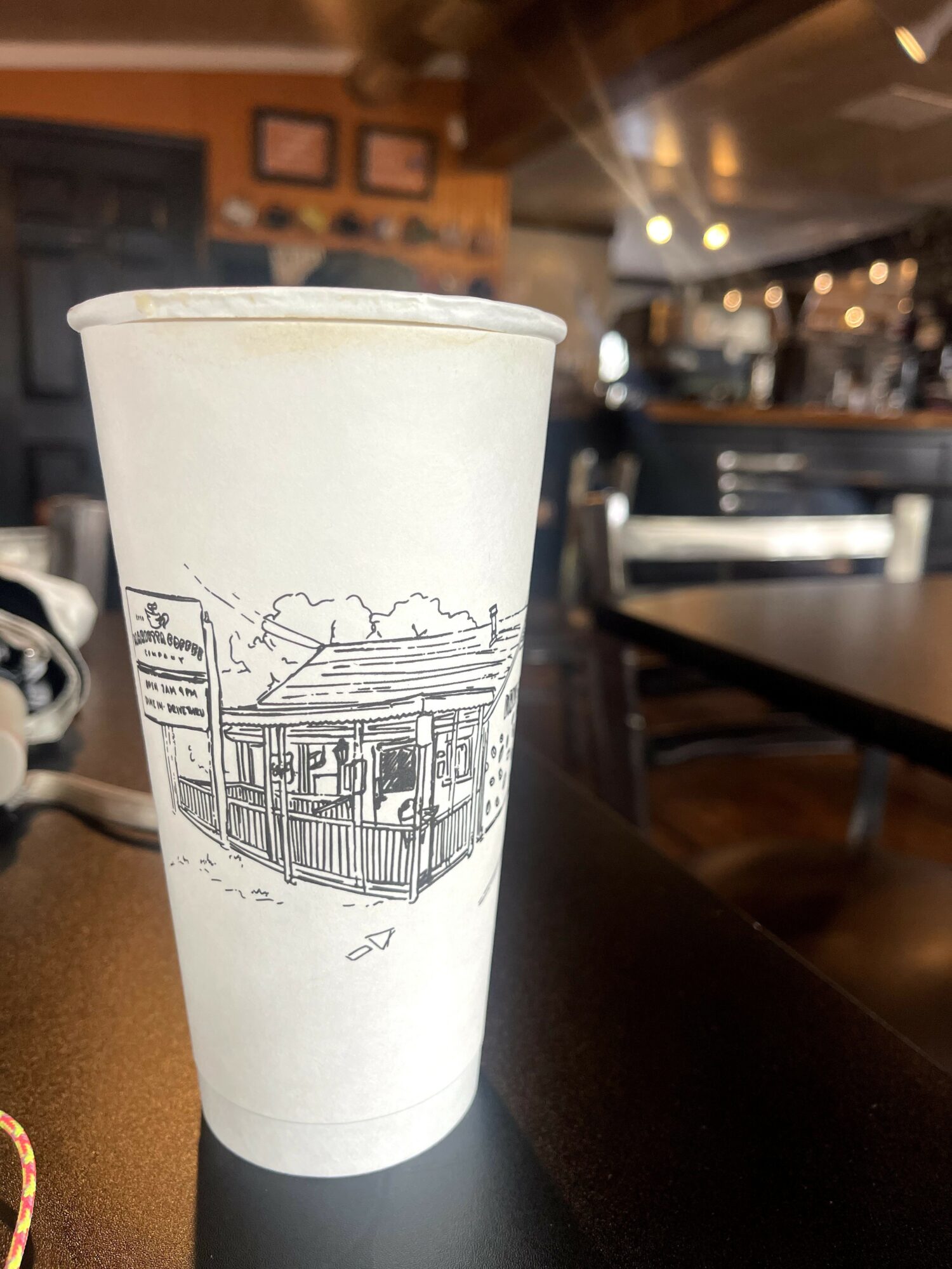 A white paper cup with a line drawing of a building and trees, placed on a table in a cafe.