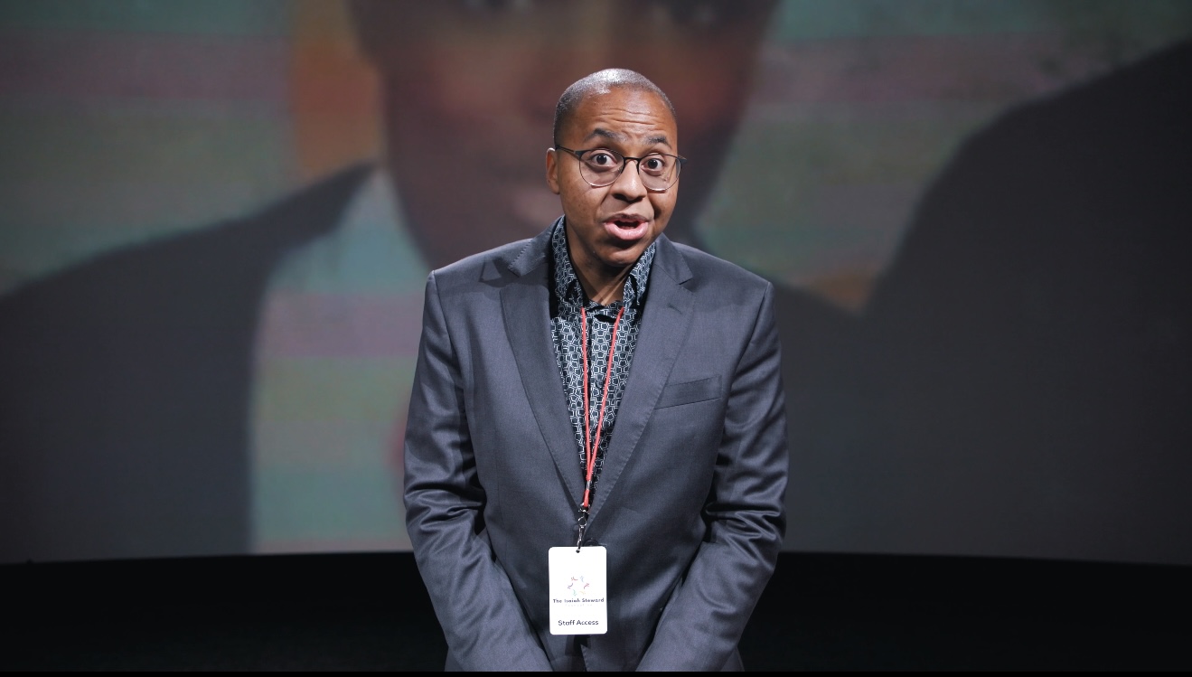 Person speaking on stage, wearing a suit and glasses, with a large screen behind them displaying a blurred image.