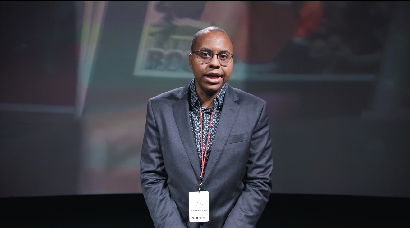 Man in a suit with glasses standing in front of a dark background.