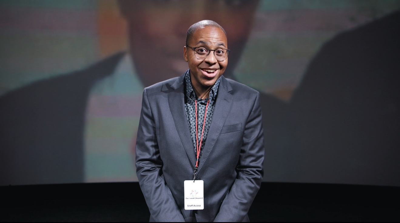 Person in a suit smiling, standing in front of a dark background, wearing a lanyard with an ID badge.