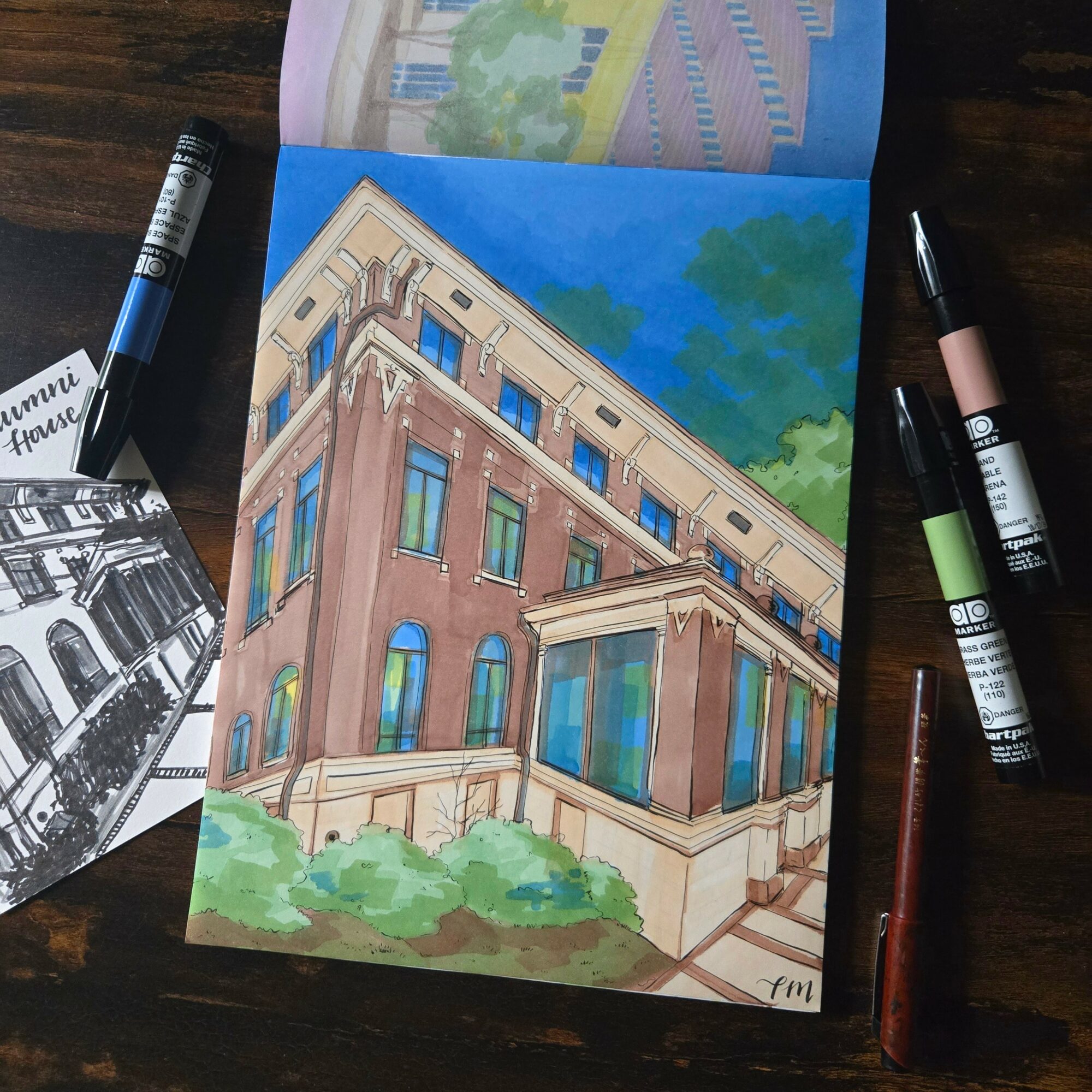 Colorful painting of a large building with columns, windows, and greenery in front, on a wooden table with pens nearby.