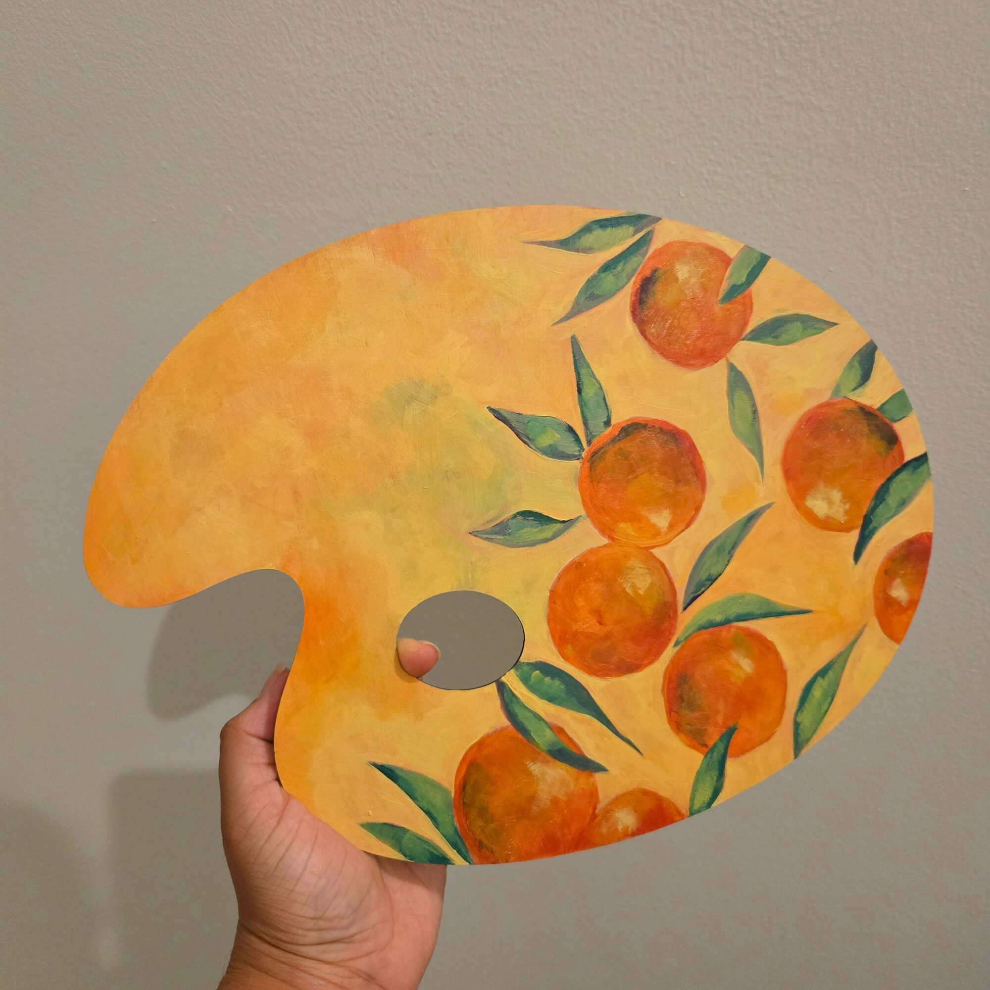 Hand holding a painted palette with oranges and green leaves, against a plain wall background.