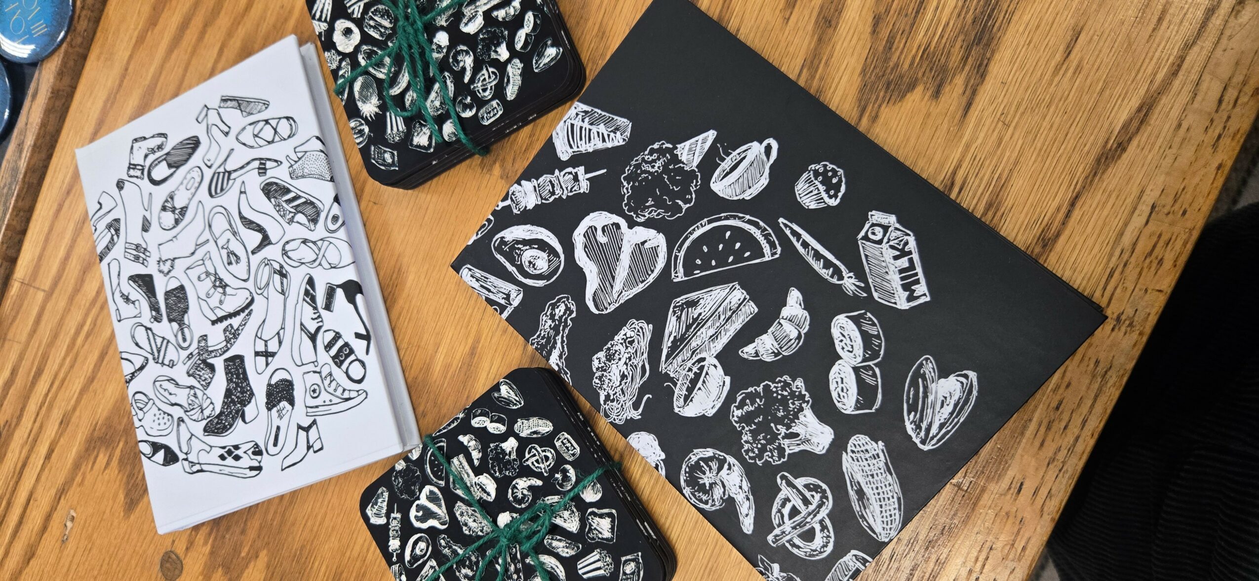 Black and white drawings of various food items on black paper, with a white sheet of similar drawings nearby, on a wooden surface.