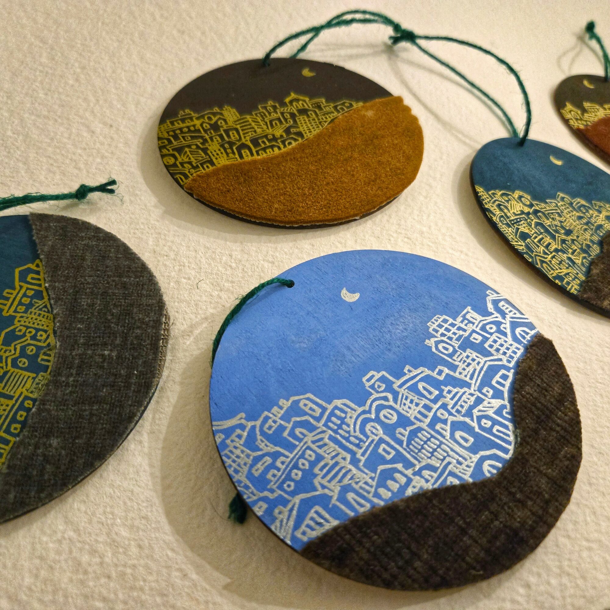 Colorful circular fabric ornaments with cityscape and moon designs, hanging on strings against a textured wall.