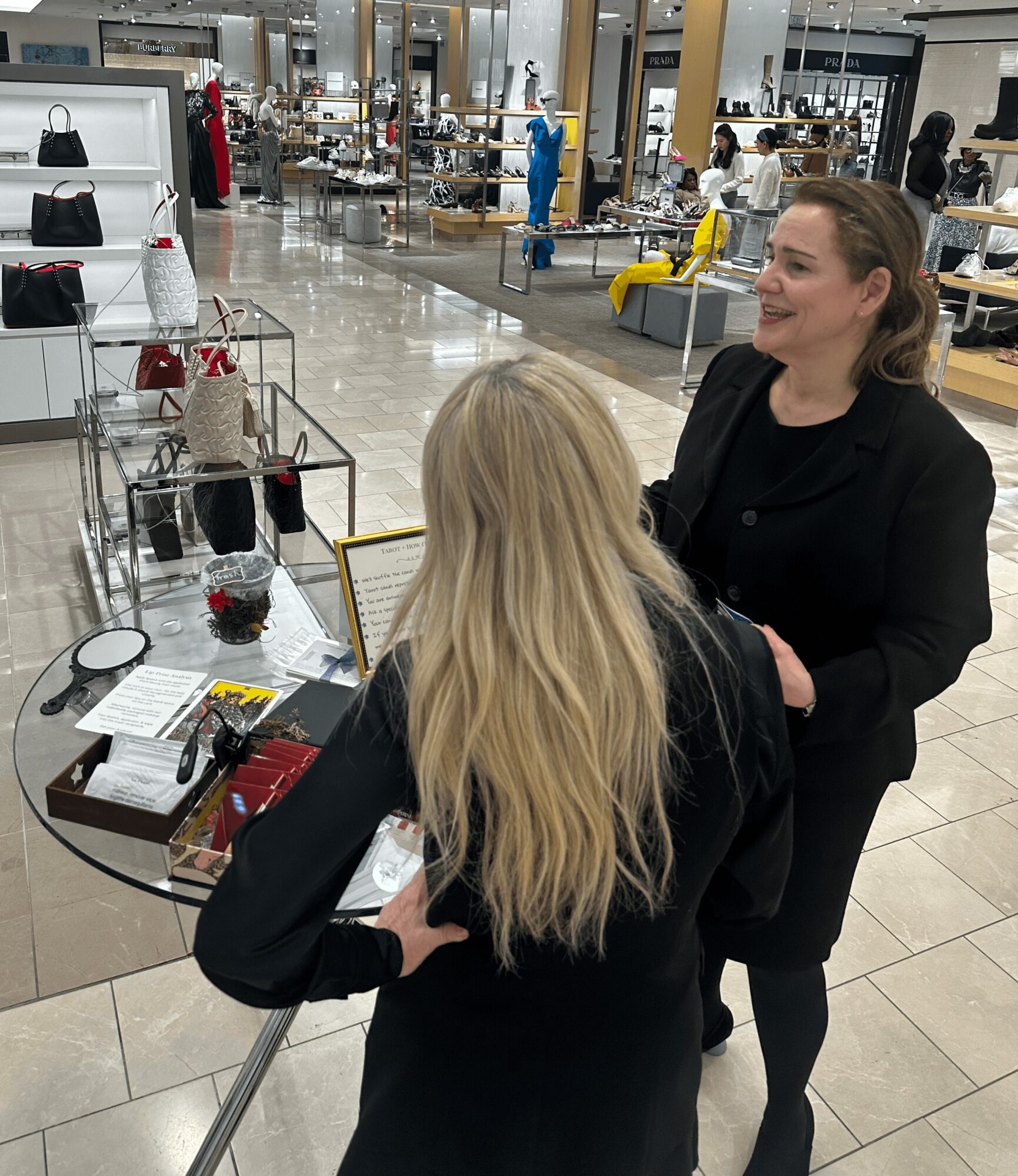 Two women talking in a shopping mall, one with long blonde hair and the other with dark hair, near a display table.