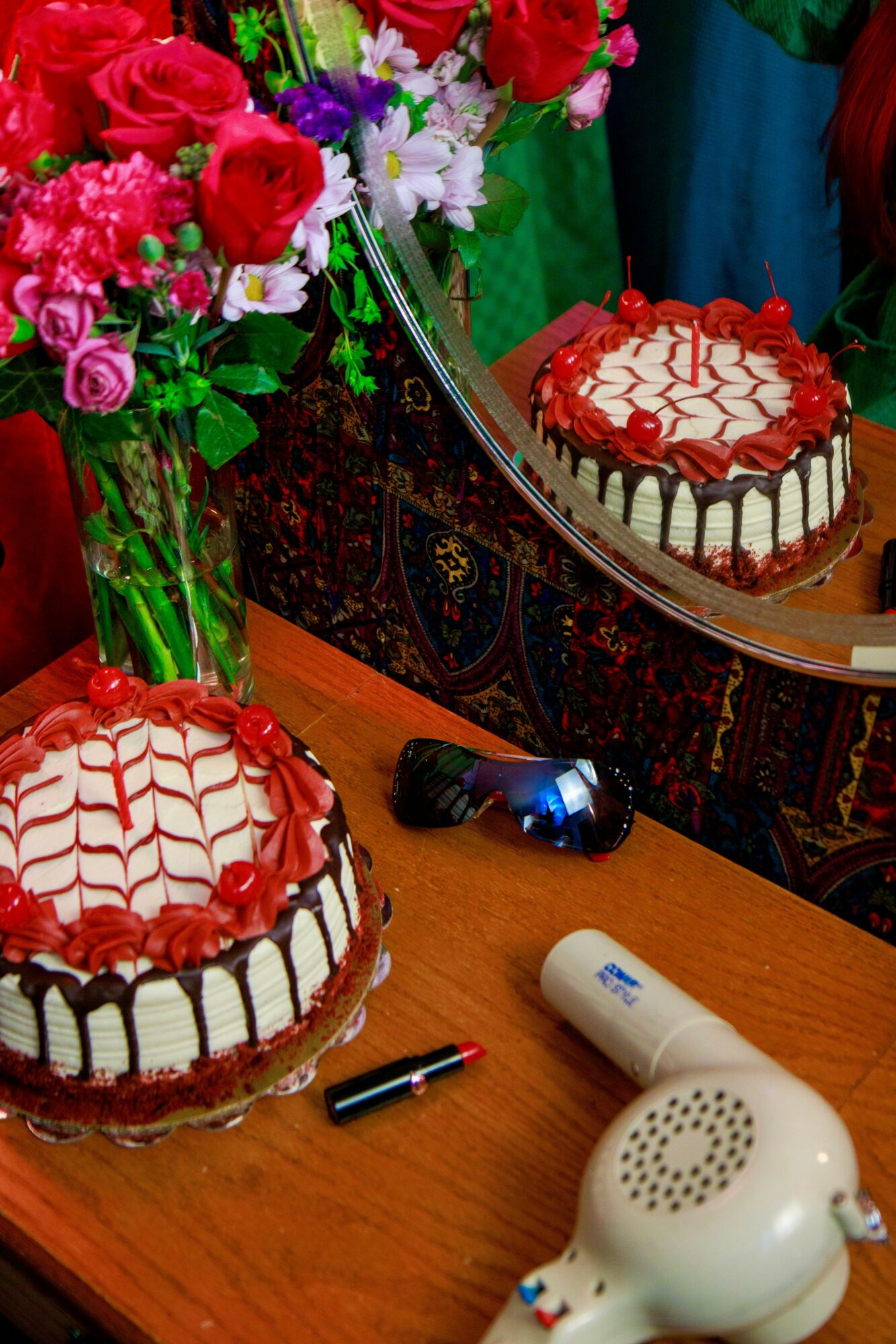 Two decorated cakes on a table with a bouquet of flowers, a mirror, and personal items, including a hairdryer and a pen.