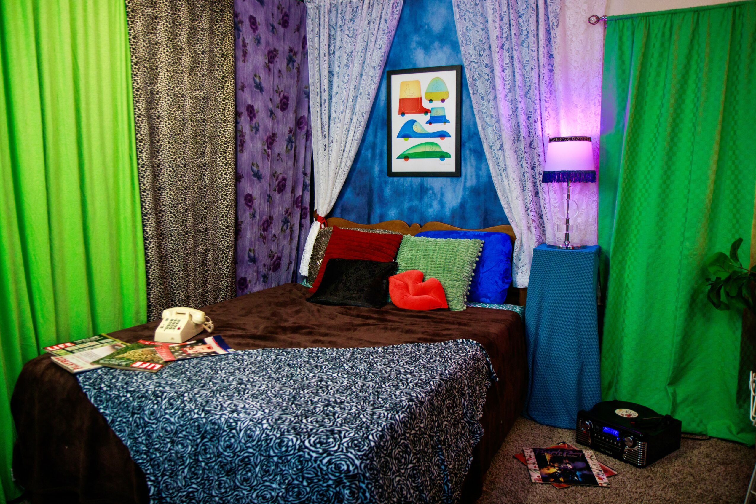 Bedroom with colorful curtains, bed with pillows, artwork, and a nightstand with a phone and magazines.